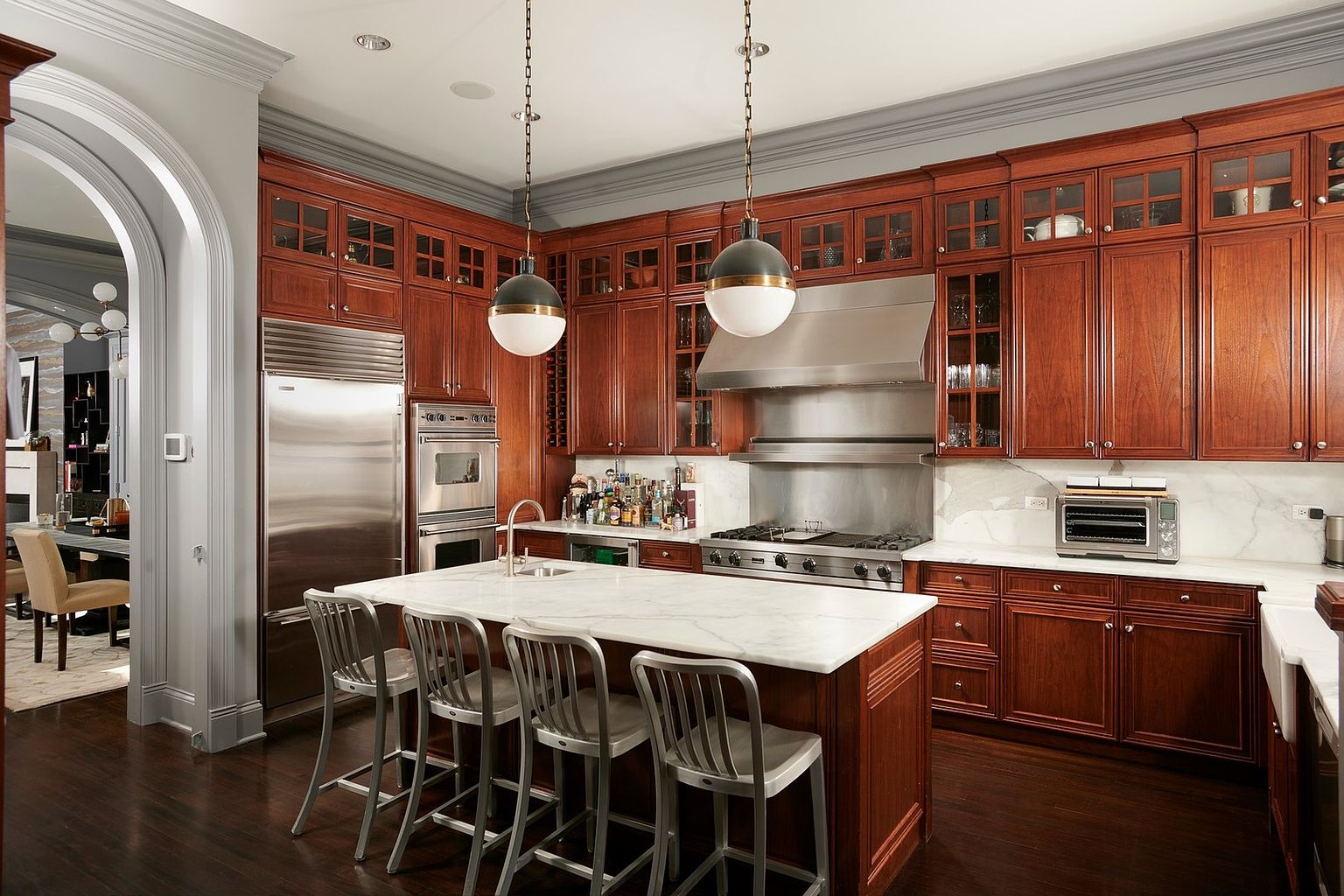 This elegant, traditional-style kitchen features rich cherry wood cabinetry, a large marble-topped center island, and high-end stainless steel appliances. The space is anchored by two sophisticated pendant lights hanging over the island, while an arched doorway leads into an adjacent dining area. The combination of dark hardwood floors, marble countertops, and classic crown molding creates a warm, luxurious, and inviting atmosphere.
