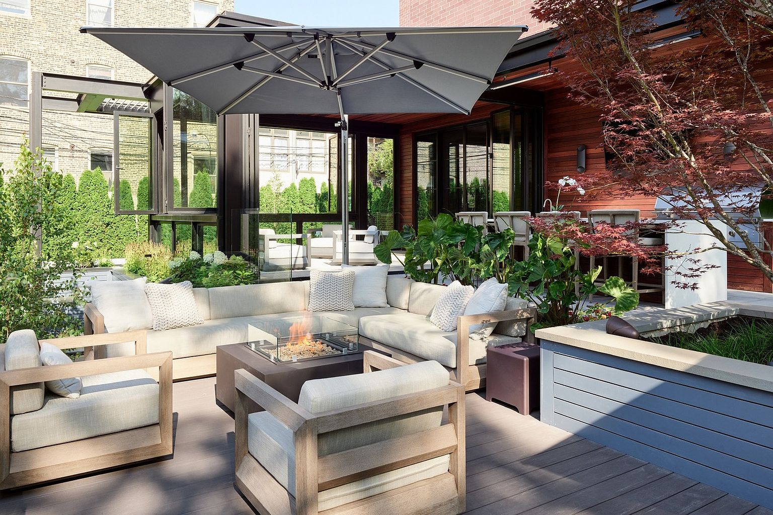 This sophisticated outdoor living space features a spacious wooden deck furnished with a modern sectional sofa, matching armchairs, and a central fire pit. The area is shaded by a large, contemporary cantilever umbrella and is surrounded by lush greenery, including a vibrant Japanese maple tree. The seamless transition between the indoor living area and the outdoor deck creates an inviting, high-end atmosphere perfect for entertaining.