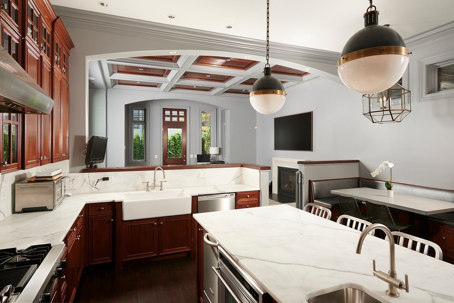 This elegant, traditional-style kitchen features rich cherry wood cabinetry, white marble countertops, and a classic farmhouse sink. The space is anchored by a large central island and flows seamlessly into an adjacent dining area with a built-in banquette, all illuminated by sophisticated pendant lighting. The perspective captures the open-concept layout, highlighting the coffered ceiling and the transition toward the living area with a fireplace.