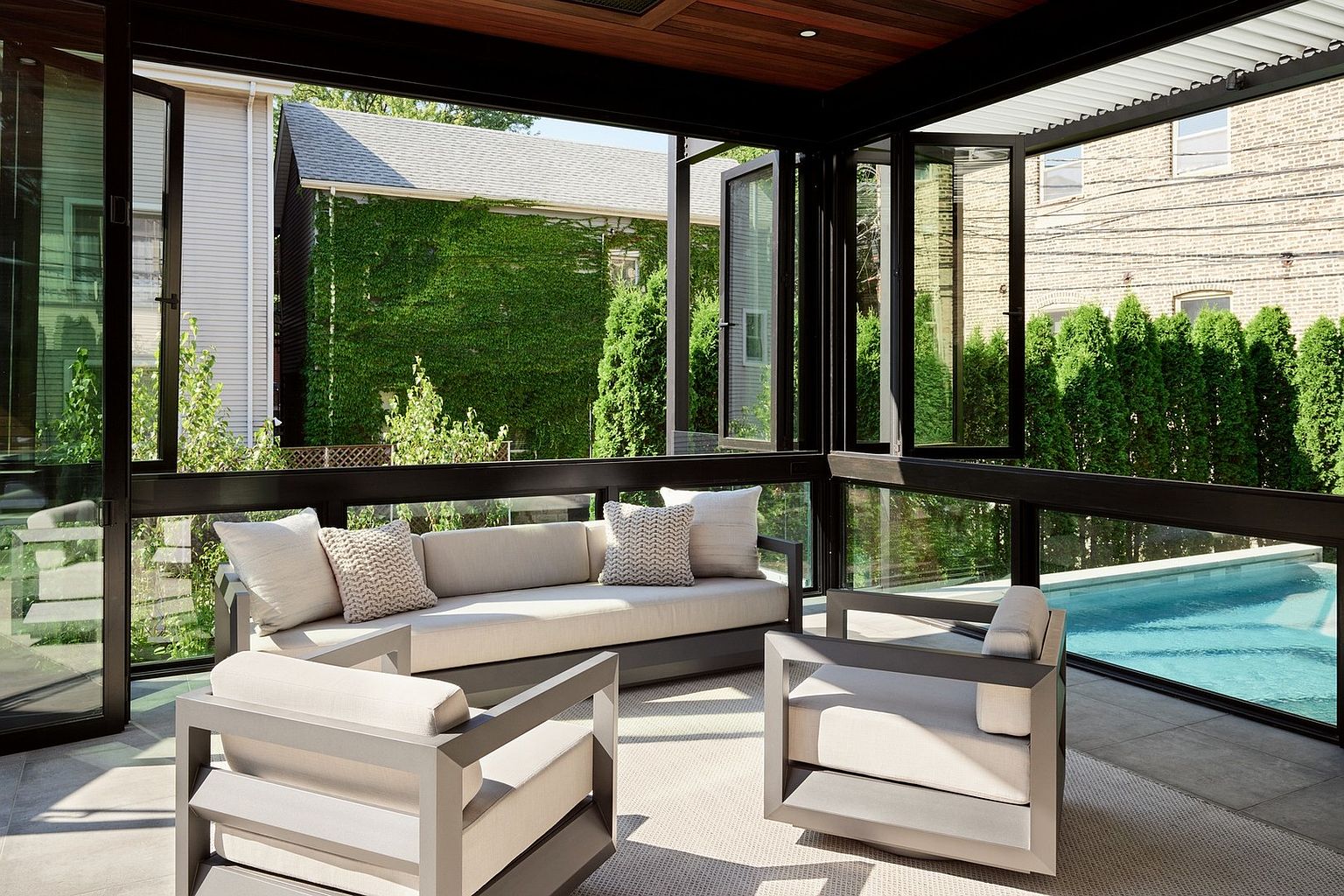 This sophisticated sunroom features floor-to-ceiling glass walls that seamlessly blend indoor comfort with the outdoor pool area. The space is furnished with a modern, neutral-toned sofa and matching armchairs, all set atop a textured area rug. The open-concept design, highlighted by dark metal framing and a warm wood-paneled ceiling, creates an airy and luxurious atmosphere perfect for relaxation.