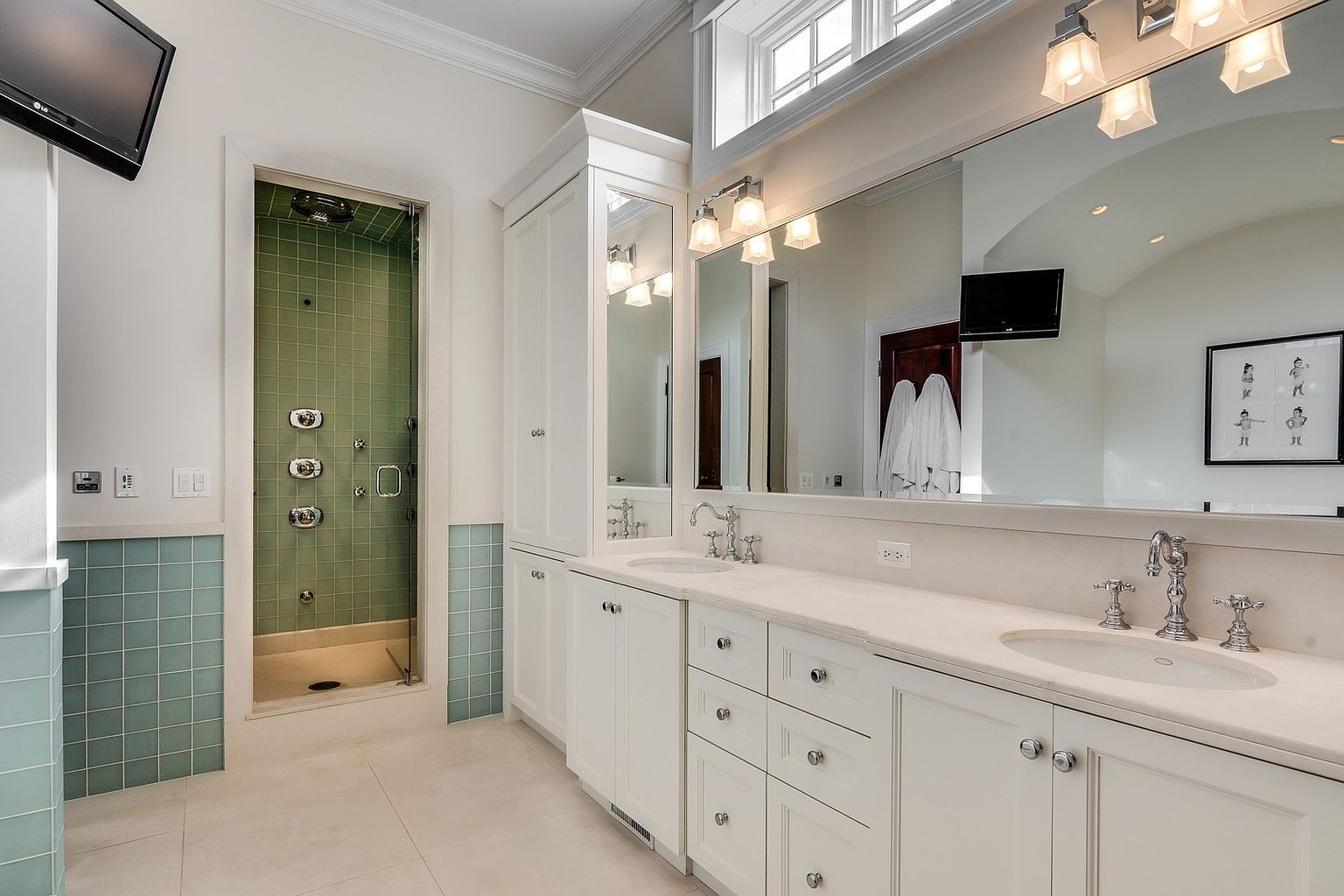 This elegant primary bathroom features a double vanity with white cabinetry, marble countertops, and polished chrome fixtures. A glass-enclosed shower with distinctive green subway tiles adds a pop of color, while a large mirror and transom window create a bright, airy atmosphere. The space is well-appointed with high-end finishes and a clean, sophisticated design.