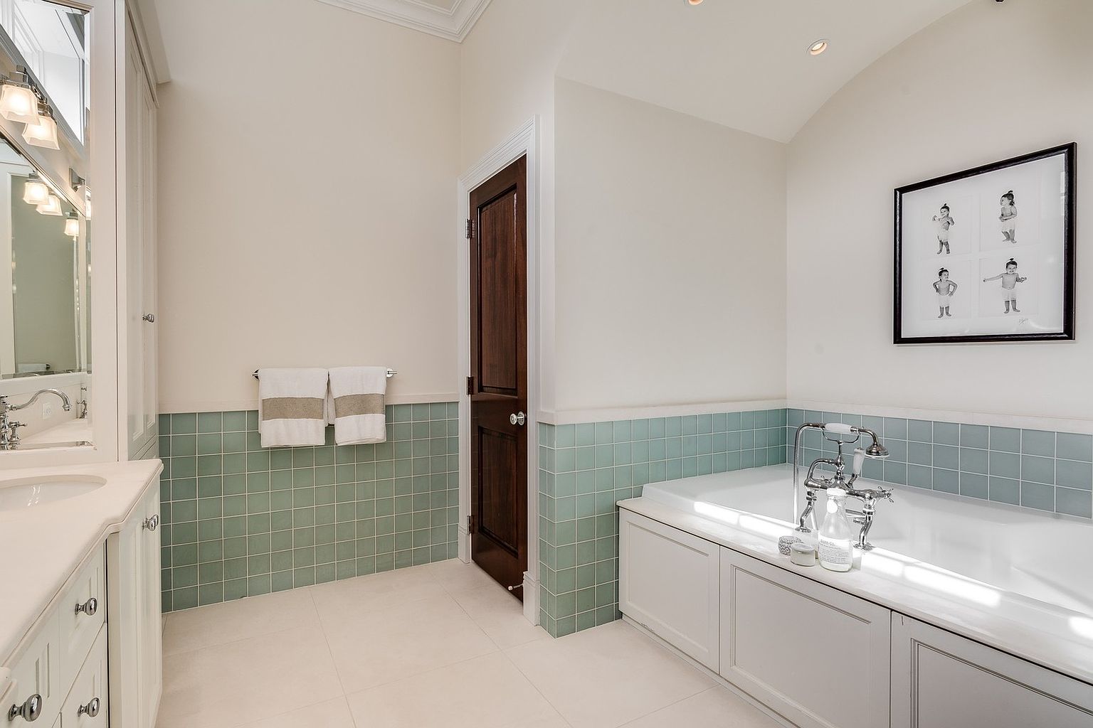 This bright and airy primary bathroom features a classic design with a deep soaking tub, a white vanity, and charming sage green square wall tiles. The space is accented by a dark wood door, polished chrome fixtures, and a framed piece of art, creating a clean and inviting atmosphere. The perspective is a wide-angle shot that captures the functional layout and elegant aesthetic of the room.