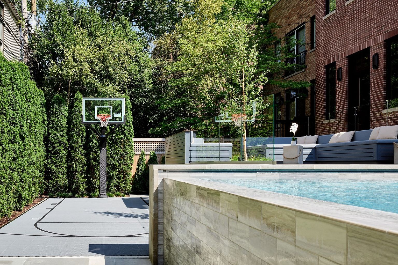 This sophisticated outdoor living space features a sleek, modern swimming pool with light-toned stone coping and a matching tiled wall. Adjacent to the pool is a dedicated basketball court area and a comfortable lounge seating arrangement, all framed by lush green privacy hedges and mature trees. The scene captures a high-end, resort-style backyard designed for both recreation and relaxation.