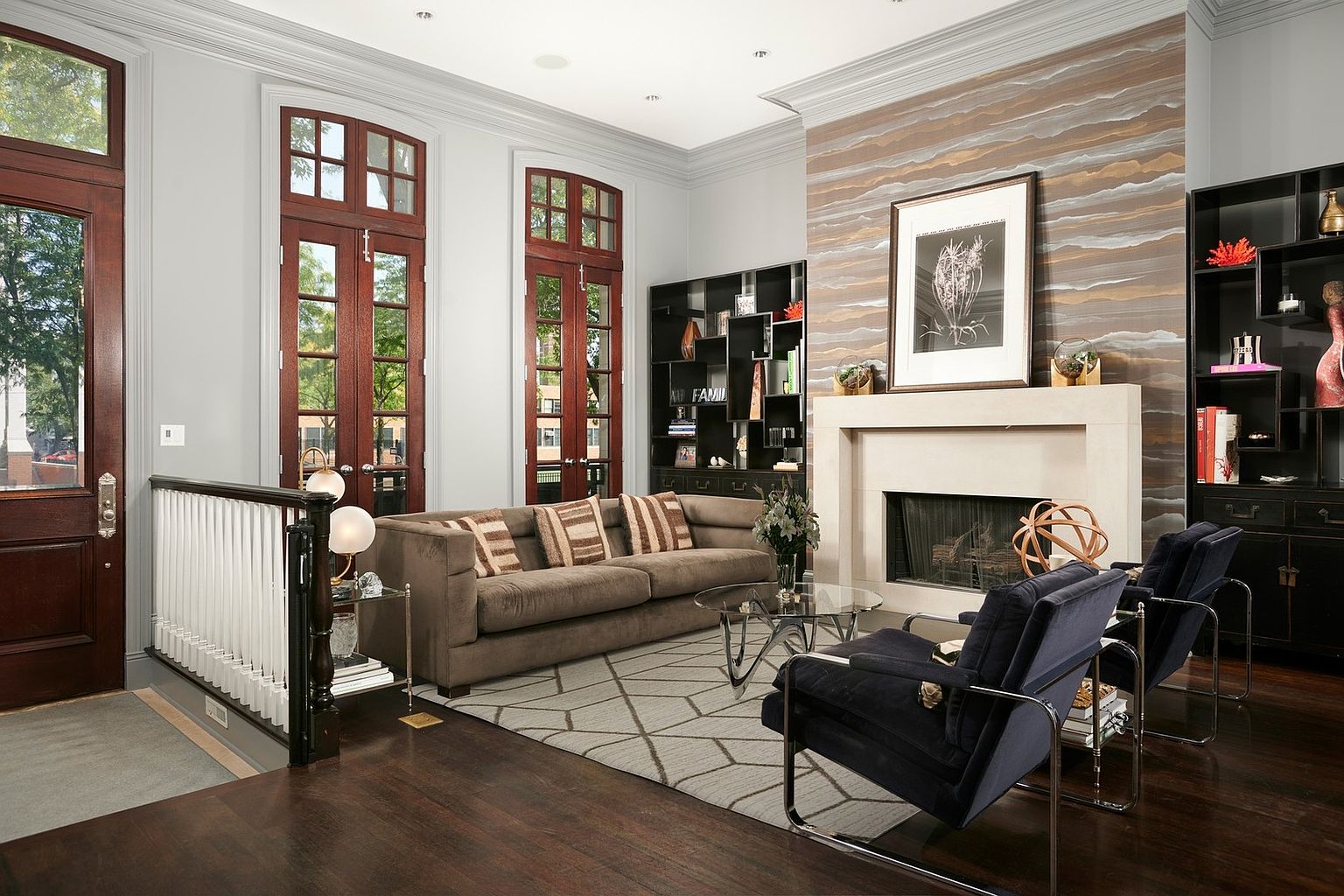 This sophisticated living room features a warm, inviting color palette with a plush brown sofa and two navy blue velvet armchairs arranged around a glass-topped coffee table. The focal point is a striking fireplace wall adorned with textured, horizontal-striped wallpaper and a large framed botanical print, flanked by sleek black built-in shelving. The room is bathed in natural light from tall, dark-wood-framed French doors, creating an elegant and polished atmosphere perfect for high-end residential living.