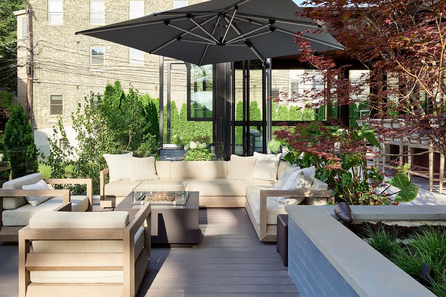 This inviting outdoor living space features a modern, neutral-toned sectional sofa and matching armchairs arranged around a sleek, square fire pit on a composite deck. The area is shaded by a large, contemporary cantilever umbrella and surrounded by lush greenery, including a Japanese maple, creating a private and serene atmosphere. The perspective is a wide, eye-level shot that highlights the seamless transition between the indoor living area and the well-appointed outdoor lounge.