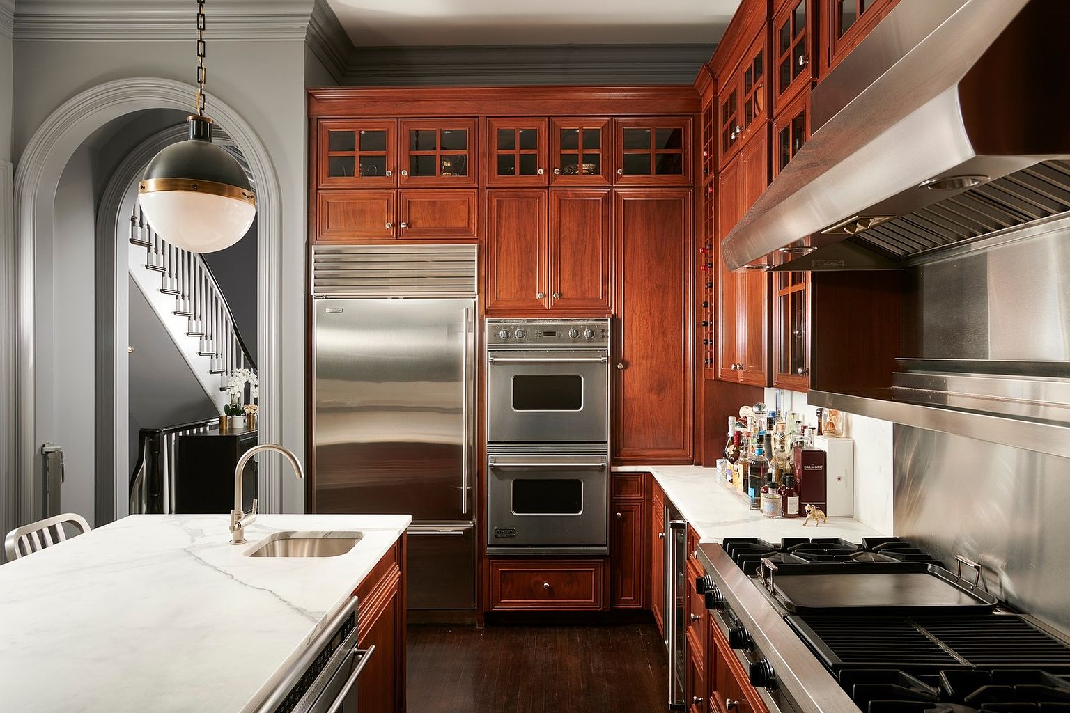 This elegant, high-end kitchen features rich cherry wood cabinetry, professional-grade stainless steel appliances, and pristine white marble countertops. The space is anchored by a large central island and flows seamlessly into an arched entryway that reveals a grand staircase, highlighting a sophisticated and classic architectural style. The perspective is a wide-angle shot from the kitchen island, emphasizing the luxury finishes and the open, airy layout of the home.