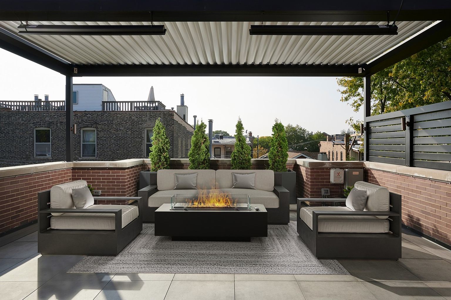 This sophisticated rooftop terrace features a modern outdoor lounge area complete with a sleek fire pit, a contemporary sofa, and two matching armchairs. The space is sheltered by a stylish louvered pergola and framed by brick walls and manicured evergreen planters, offering an urban oasis feel. The perspective is a centered, eye-level shot that highlights the inviting seating arrangement and the surrounding cityscape.