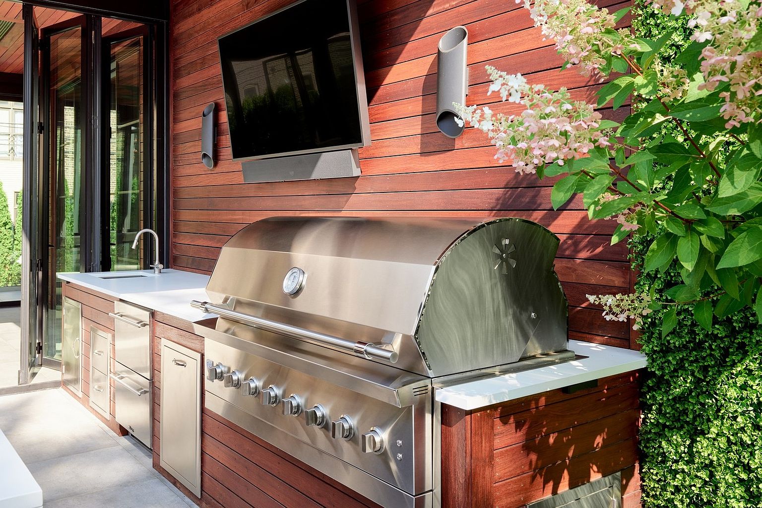 This high-end outdoor kitchen features a sleek stainless steel grill and matching cabinetry set against a warm, horizontal wood-slatted wall. A wall-mounted television and integrated sound system enhance the entertainment potential, while a lush green plant adds a vibrant, natural touch to the space. The clean lines and premium materials create a sophisticated and inviting atmosphere for outdoor dining and relaxation.