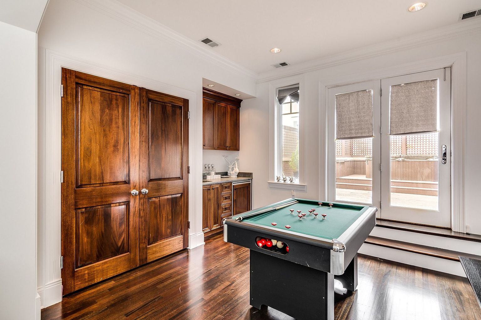 This interior space functions as a game room, featuring a central pool table with a green felt surface and a set of billiard balls. To the left, a set of large, rich-toned wooden double doors adds a classic touch, while a built-in wet bar with dark cabinetry sits in the corner. The room is brightened by natural light streaming through a tall window and glass French doors that lead out to a deck, creating an inviting area for entertainment.