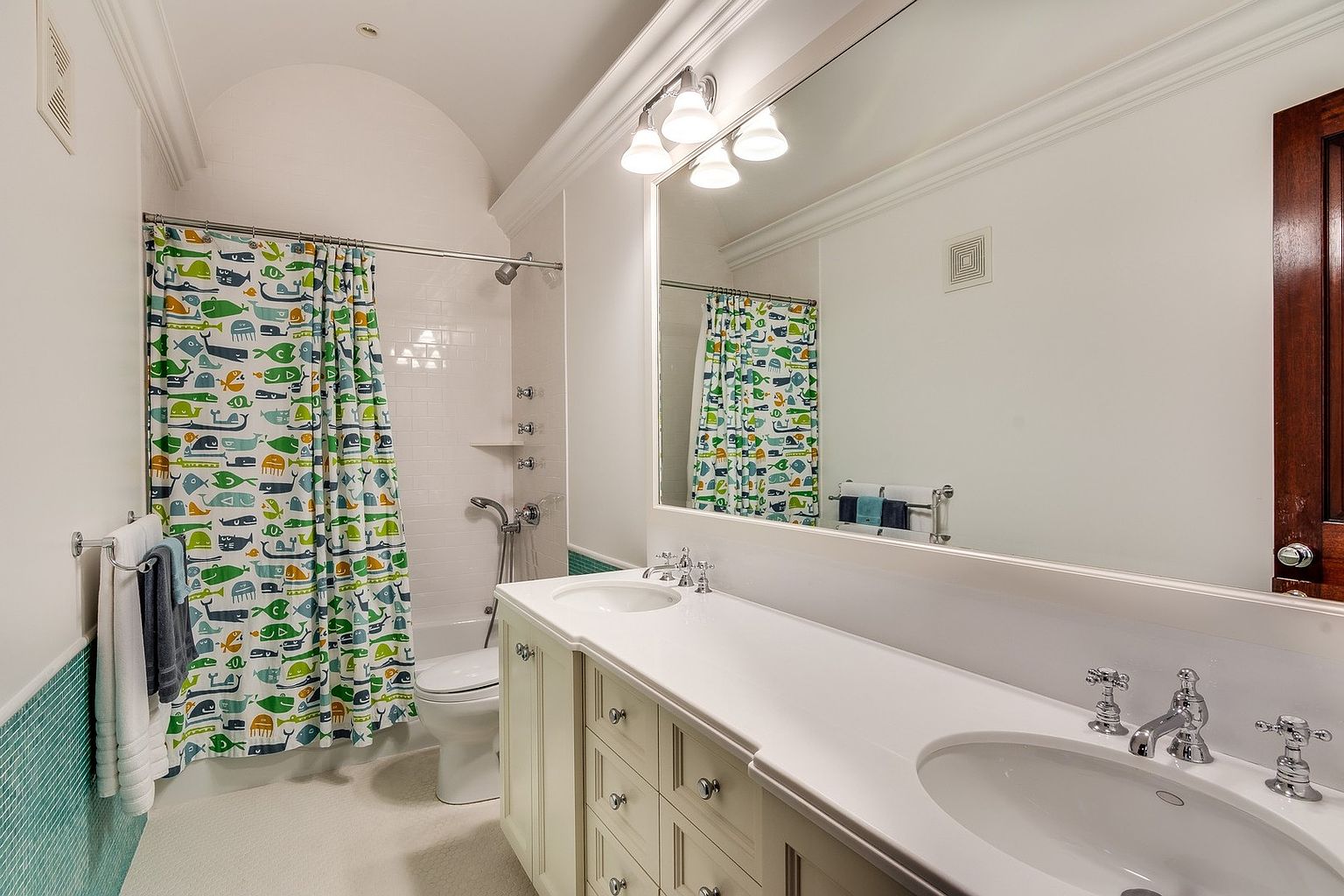 This bright and playful bathroom features a double vanity with a white countertop and light-colored cabinetry, complemented by a large wall-to-wall mirror. A whimsical fish-patterned shower curtain adds a pop of color to the space, which also includes a white subway-tiled shower area and teal accent tiling along the lower walls. The room is well-lit by a multi-bulb vanity light fixture, creating a clean and inviting atmosphere suitable for a guest or children's bathroom.