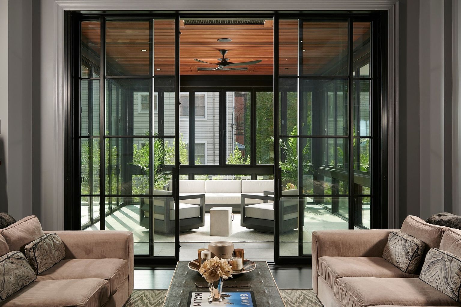 This sophisticated living room features plush, neutral-toned sofas framing a central coffee table, creating an inviting and elegant atmosphere. The space is anchored by a massive, floor-to-ceiling black-framed glass wall that seamlessly connects the interior to a covered outdoor patio, which is furnished with modern lounge seating and a wood-paneled ceiling. The perspective is perfectly centered, emphasizing the architectural harmony between the refined indoor living area and the serene, light-filled outdoor retreat.