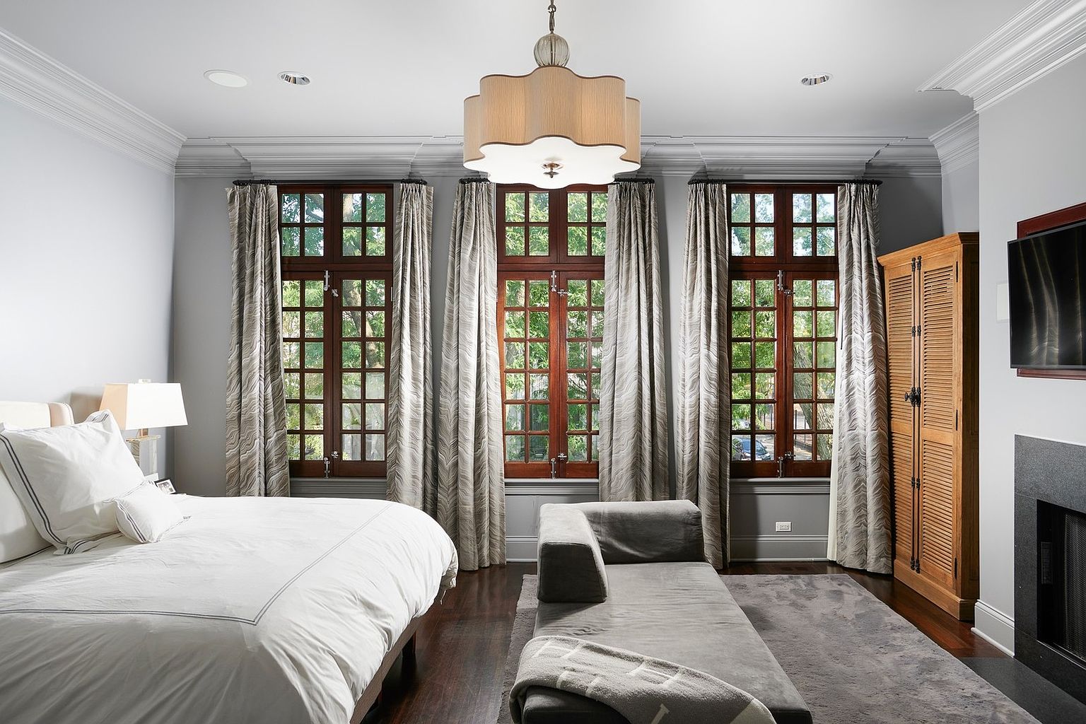 This elegant primary bedroom features a sophisticated neutral color palette, highlighted by dark hardwood floors and three large, wood-framed windows dressed in patterned floor-to-ceiling curtains. A plush grey chaise lounge sits atop a matching area rug at the foot of a neatly made bed, while a tall wooden armoire and a sleek fireplace add warmth and character to the space. The room is anchored by a distinctive, scalloped-edge chandelier, creating a refined and tranquil atmosphere.
