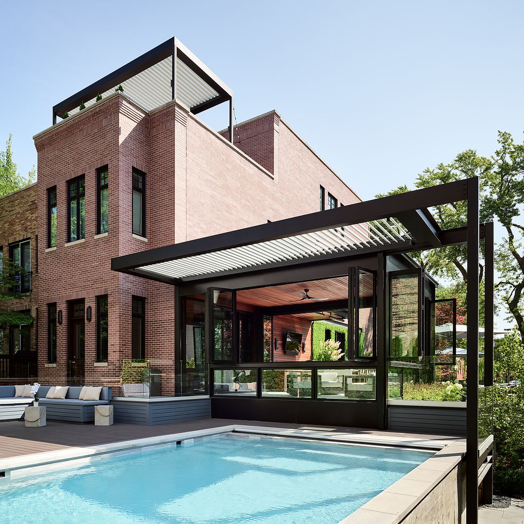 This sophisticated outdoor living space features a modern, rectangular swimming pool set against a striking brick residence. A sleek, black-framed pergola structure extends from the house, housing an indoor-outdoor lounge area with folding glass doors that seamlessly blend the interior and exterior environments. The scene exudes luxury and contemporary design, perfect for high-end entertaining.
