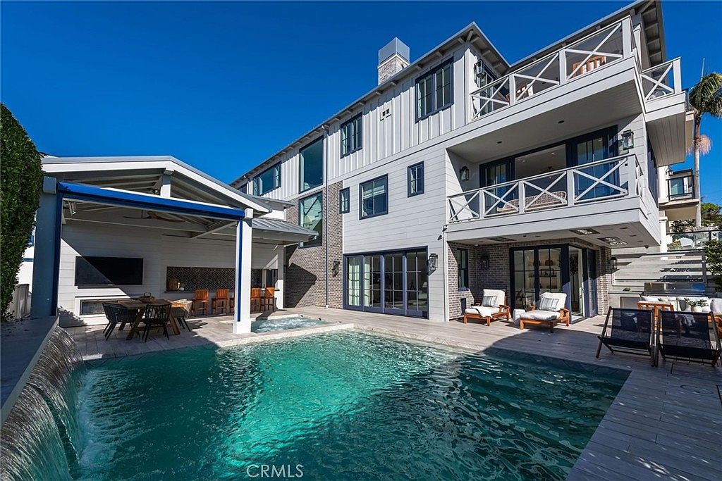 This rear exterior view showcases a luxurious multi-level home with a modern aesthetic, featuring a combination of white siding and brick accents. A sparkling swimming pool with an integrated spa and a cascading water feature serves as the centerpiece of the backyard, complemented by a covered outdoor dining area and multiple balconies. The scene is captured from a low angle, emphasizing the expansive outdoor living space and the seamless transition between the indoor and outdoor areas.