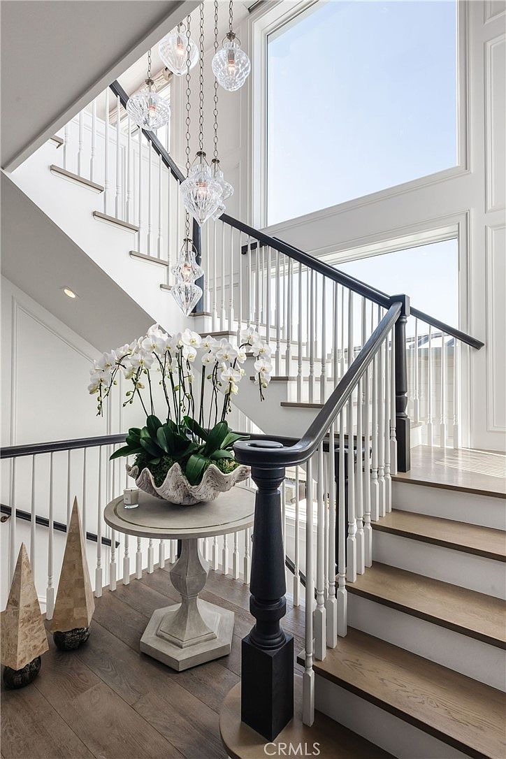 This elegant entryway features a grand staircase with dark wood handrails and white balusters, leading up to a landing bathed in natural light from large windows. A round pedestal table sits at the base of the stairs, topped with a large, decorative shell-shaped bowl filled with white orchids. A cluster of glass pendant lights hangs from the ceiling, adding a sophisticated, modern touch to the bright and airy space.