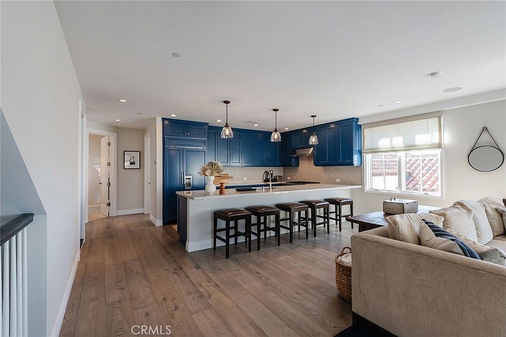 This open-concept kitchen features striking deep blue cabinetry contrasted by a light-toned island and light wood flooring. The space is illuminated by three pendant lights hanging over the island, which provides seating for four, and includes a view into an adjacent living area. The perspective is from the living room looking toward the kitchen, highlighting the seamless flow between the two living spaces.