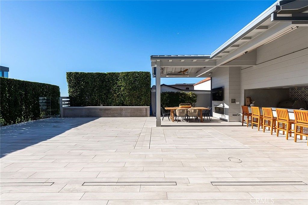 This expansive outdoor patio features light-toned, wood-look plank flooring and a covered dining area sheltered by a modern pergola. The space includes a built-in outdoor kitchen with bar-style seating and a separate dining table, all set against a backdrop of tall, manicured privacy hedges. The clean lines and neutral color palette create a sophisticated, contemporary atmosphere perfect for al fresco entertaining.