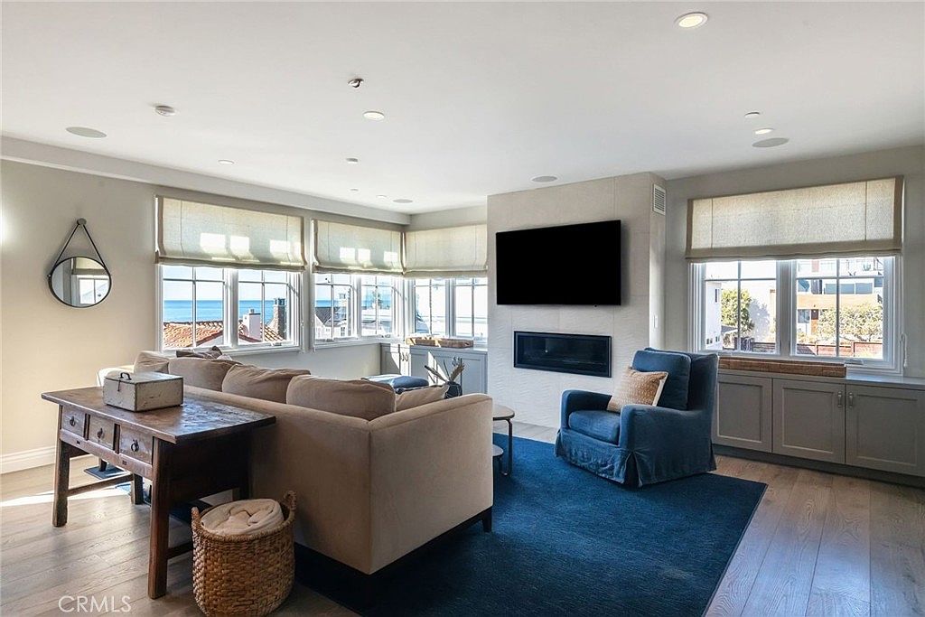 This bright and airy living room features a neutral color palette with light wood flooring and a large blue area rug. The space is anchored by a comfortable sofa and a blue armchair, centered around a wall-mounted television above a linear fireplace. Large windows with light-colored shades offer expansive views of the ocean, creating a serene and inviting coastal atmosphere.