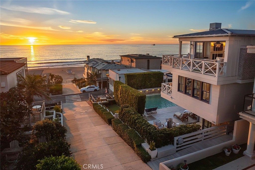 This elevated perspective captures a multi-story coastal residence at sunset, showcasing its prime location near the beach. The property features a private pool area enclosed by tall, manicured hedges, a spacious deck, and multiple balconies that offer expansive ocean views. The scene highlights the seamless integration of indoor-outdoor living, with the golden hour light casting a warm glow over the neighborhood and the horizon.
