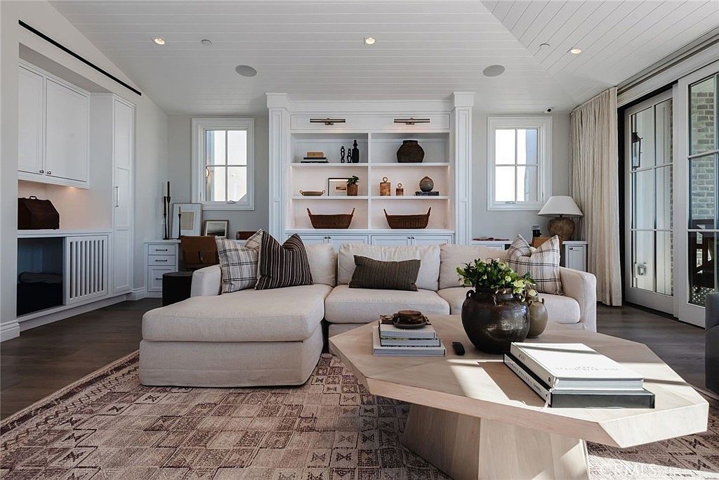 This bright and airy living room features a large, neutral-toned sectional sofa centered around a unique hexagonal light wood coffee table. A built-in shelving unit with integrated lighting serves as a focal point, displaying curated decor, while a patterned area rug adds texture to the dark wood flooring. The space is framed by tall windows and glass doors that allow for ample natural light, creating a sophisticated and inviting atmosphere.