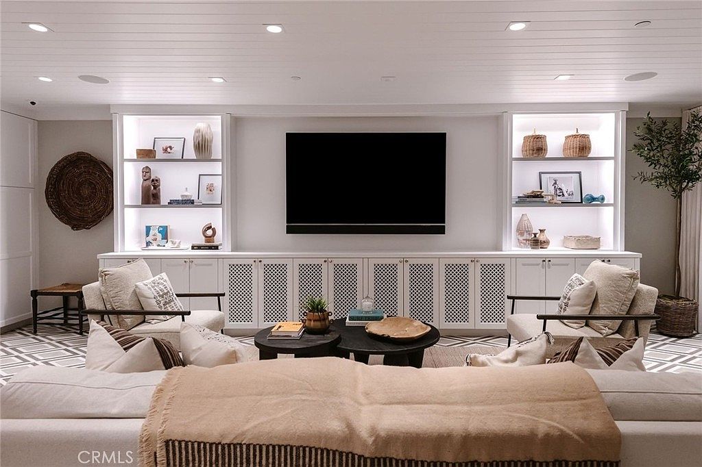 This inviting living room features a symmetrical built-in media wall with illuminated shelving flanking a central television. The space is furnished with two comfortable armchairs and a large sectional, centered around a pair of round coffee tables. The room is finished with a light-toned, paneled ceiling and a patterned area rug, creating a warm and cohesive aesthetic.