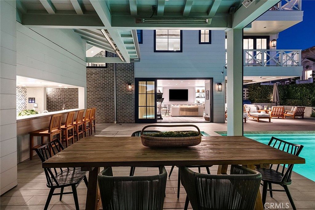 This expansive covered outdoor living space features a large wooden dining table in the foreground and a built-in bar area with high stools to the left. Beyond the covered patio, a swimming pool glows with blue light, and a transition into the indoor living room is visible through wide-opening glass doors. The space is designed for seamless indoor-outdoor entertaining, showcasing a sophisticated and modern coastal aesthetic.