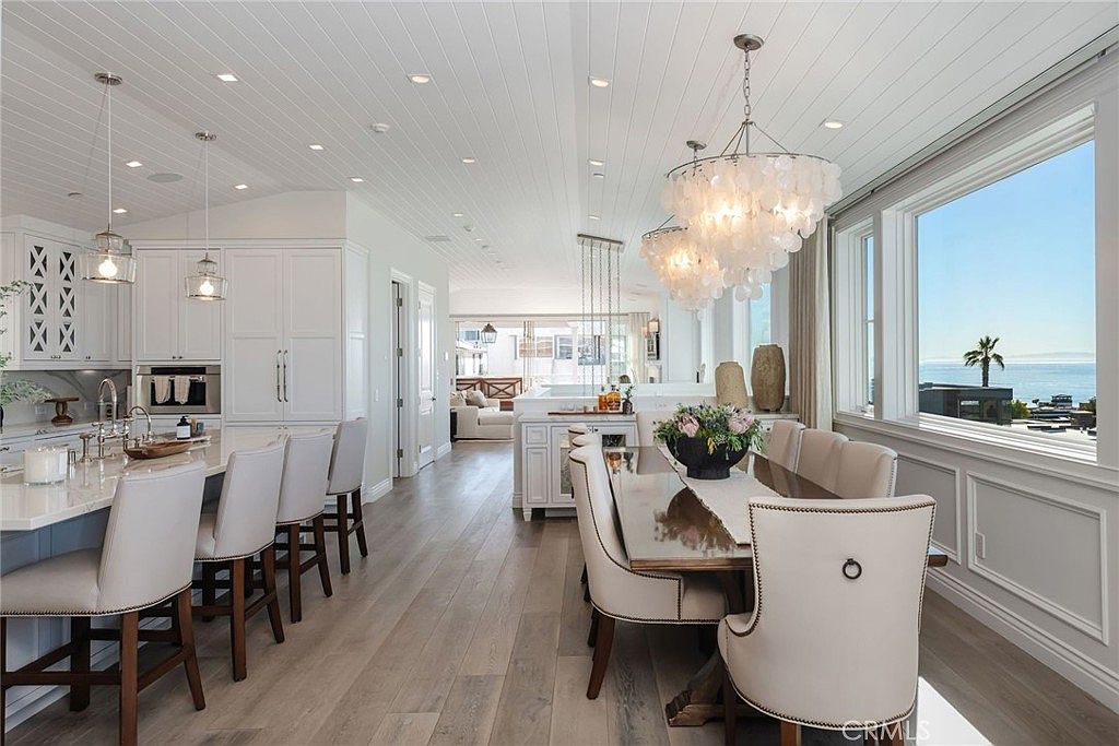 This bright, open-concept space seamlessly blends a modern kitchen with a formal dining area, characterized by light wood flooring and a white shiplap ceiling. The kitchen features a large island with bar seating, white cabinetry, and stainless steel appliances, while the adjacent dining area is anchored by a statement chandelier and large windows offering a panoramic ocean view. The overall aesthetic is airy, coastal, and sophisticated, emphasizing natural light and an expansive layout.