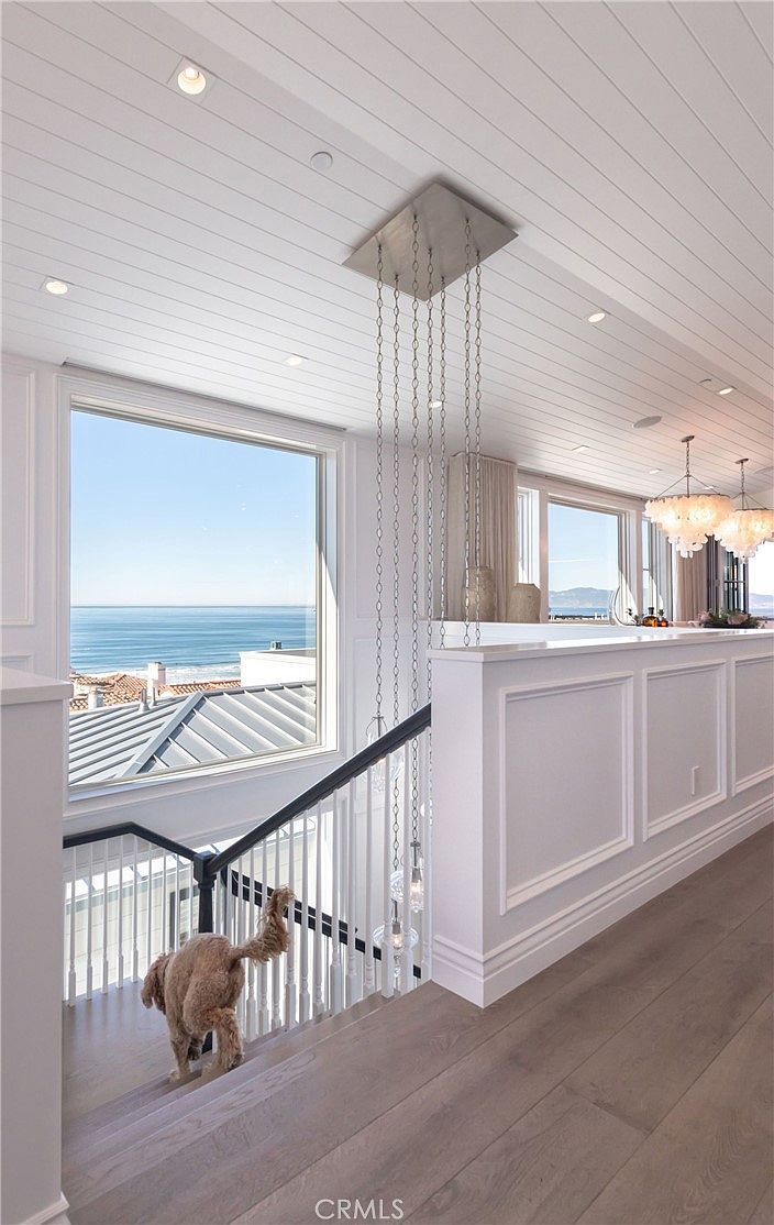 This bright, coastal-inspired staircase landing features light wood flooring and crisp white wainscoting along the walls. A dramatic, multi-tiered chain chandelier hangs from the shiplap ceiling, while large windows provide an expansive, unobstructed view of the ocean. A small dog is captured mid-step on the stairs, adding a sense of warmth and livability to this elegant, airy space.