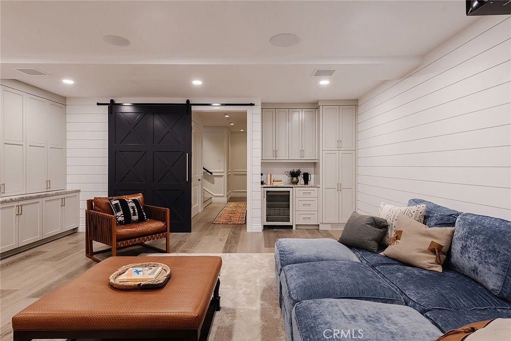 This inviting living room features a large blue sectional sofa, a leather ottoman, and a wooden armchair, all arranged on a neutral area rug. The space is accented by white shiplap walls, a dark sliding barn door, and a built-in wet bar area with a wine refrigerator. The room is illuminated by recessed lighting and offers a view into a hallway, creating a warm and cohesive living environment.