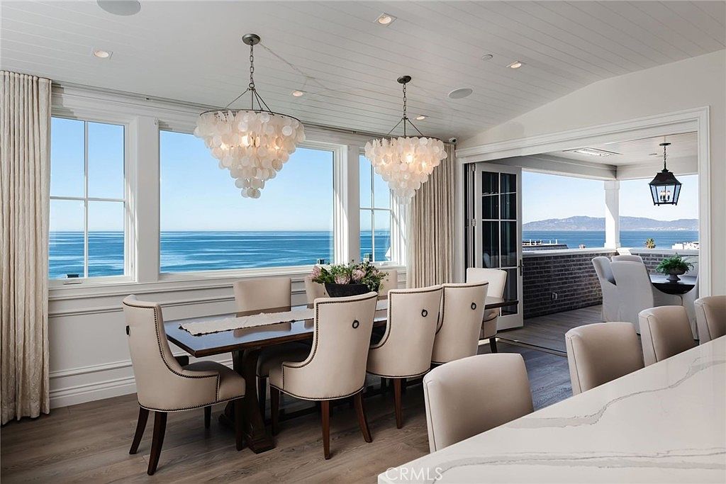 This elegant dining room features a dark wood table surrounded by upholstered chairs, set against a backdrop of expansive windows that offer panoramic ocean views. Two large, textured shell-style chandeliers hang from the white planked ceiling, adding a coastal-chic aesthetic to the space. The room seamlessly transitions to an outdoor balcony through folding glass doors, creating an airy and sophisticated atmosphere perfect for entertaining.