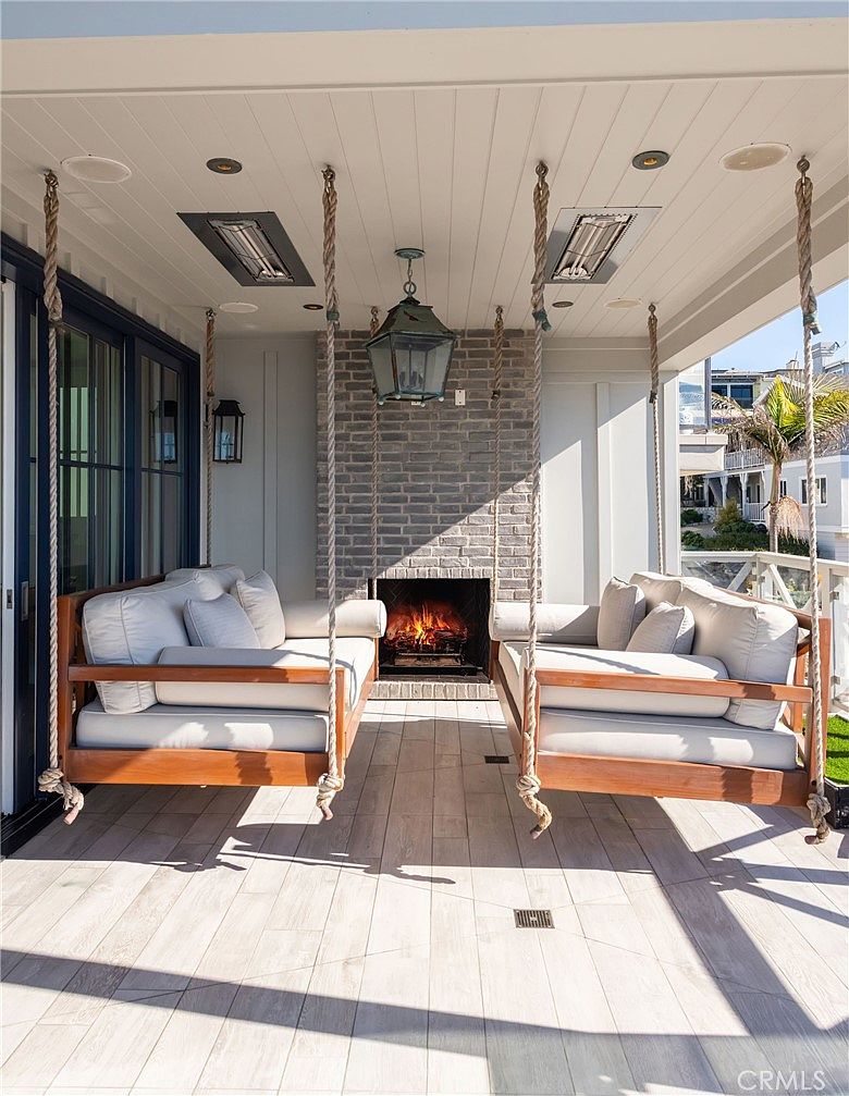 This inviting covered patio features two large, suspended wooden porch swings facing each other, creating a cozy and symmetrical seating area. A central brick fireplace serves as a focal point, adding warmth to the space, while recessed lighting and ceiling-mounted heaters provide comfort. The light-toned wood-look flooring and clean architectural lines offer a sophisticated, relaxed outdoor living experience.