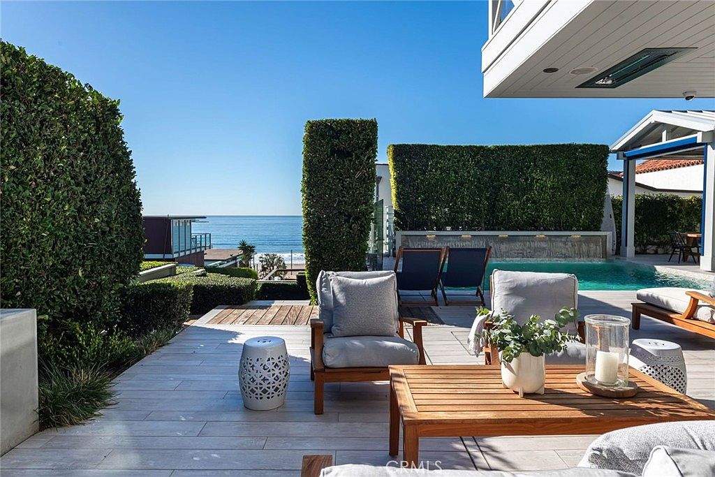 This luxurious outdoor patio space features light-toned wood-look decking and comfortable lounge seating, including armchairs and a coffee table. The area seamlessly transitions into a private swimming pool with a water feature wall, all set against a stunning, unobstructed view of the ocean. The scene is bathed in bright, natural sunlight, creating an inviting and serene atmosphere perfect for coastal living.