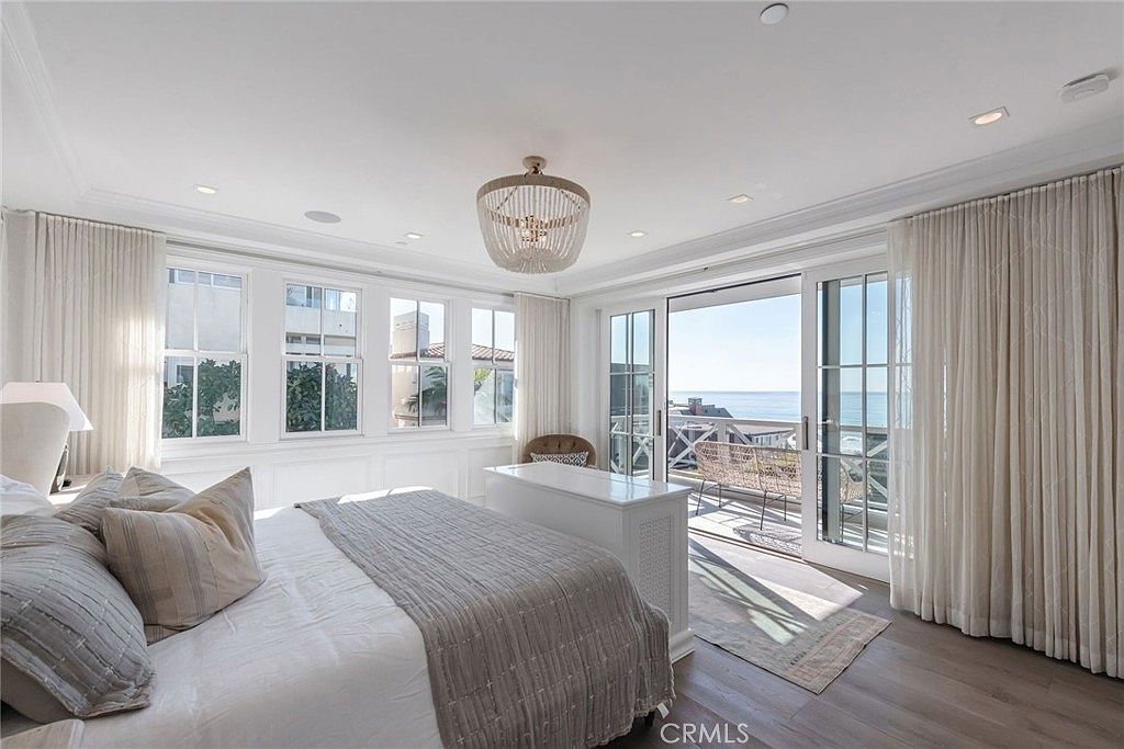 This bright and airy primary bedroom features a neutral color palette, light wood flooring, and a large bed dressed in textured linens. A beaded chandelier serves as a focal point, while a white storage console sits at the foot of the bed. Large sliding glass doors open onto a balcony, providing an expansive view of the ocean and bringing in abundant natural light.