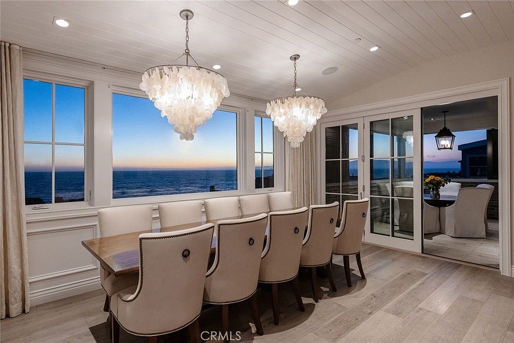 This elegant dining room features a long wooden table surrounded by upholstered chairs, positioned to take full advantage of the expansive ocean views through large windows. Two ornate, shell-inspired chandeliers hang from a white paneled ceiling, adding a sophisticated coastal aesthetic to the space. The room transitions seamlessly to an outdoor patio area through glass sliding doors, creating a bright and airy atmosphere perfect for formal dining.