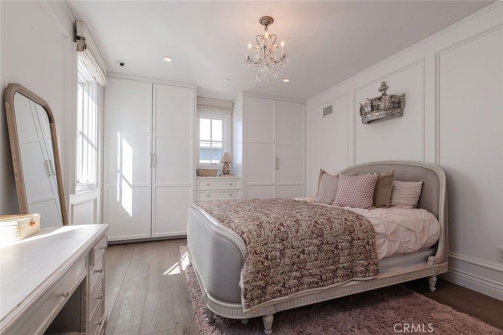 This elegant bedroom features a light-toned color palette with a classic upholstered bed frame as the focal point. The room is equipped with built-in white cabinetry for storage, a crystal chandelier, and a decorative crown wall ornament, all set against light wood flooring and a soft textured rug. A large floor mirror and a vanity area complete the space, creating a refined and cozy atmosphere.