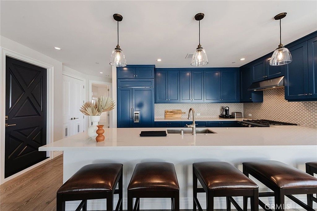 This modern kitchen features striking deep blue cabinetry contrasted by a large, light-toned island with seating for four. Three glass pendant lights hang above the island, providing focused illumination over the workspace and sink area. The space is finished with a light-colored backsplash and wood-look flooring, creating a clean and sophisticated aesthetic.