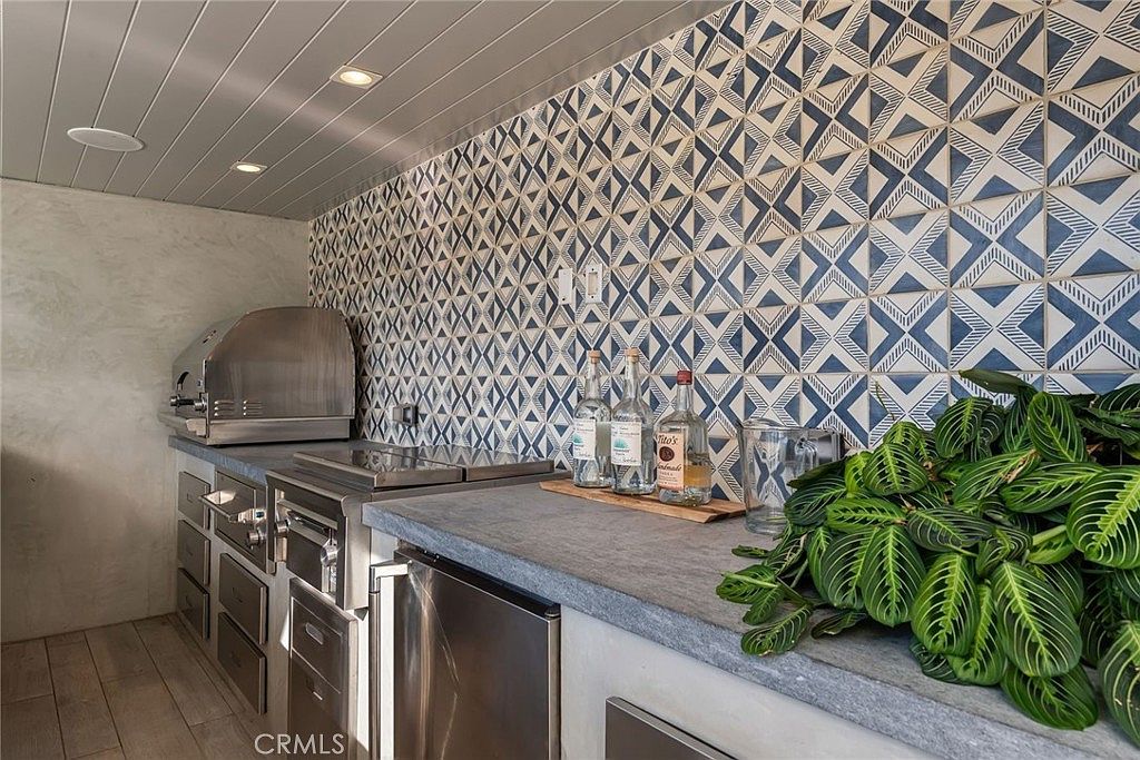This outdoor kitchen area features a stainless steel grill and a built-in cooktop set into a stone-topped counter. The wall behind the counter is adorned with a striking blue and white geometric patterned tile, adding a vibrant visual element to the space. A large potted plant sits on the right, and the area is illuminated by recessed lighting set into a paneled ceiling.