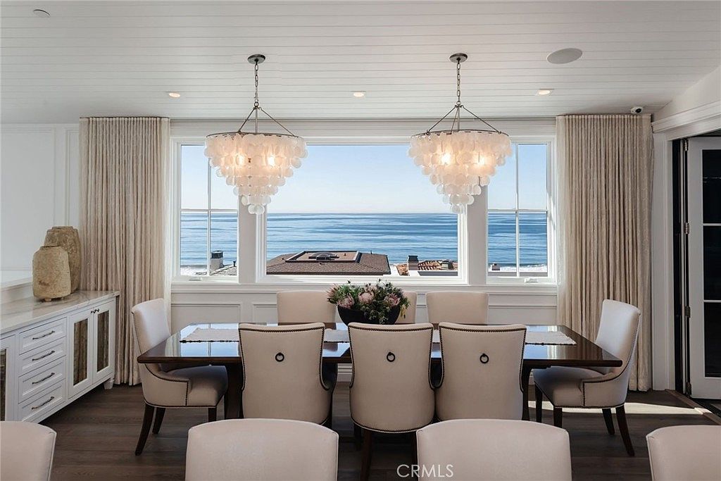 This elegant dining room features a large rectangular table surrounded by upholstered chairs, positioned to take full advantage of a panoramic ocean view through a wide window. Two ornate, shell-inspired chandeliers hang above the table, adding a coastal-chic aesthetic to the space. The room is framed by floor-length curtains and includes a built-in sideboard, creating a sophisticated and serene atmosphere perfect for formal dining.