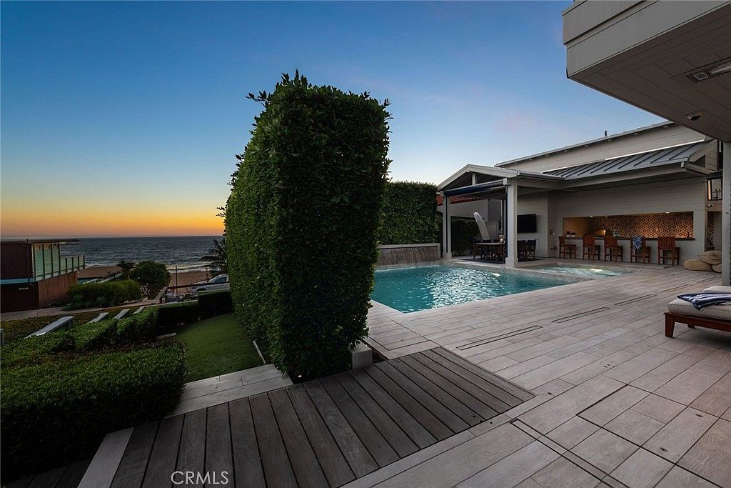 This luxurious outdoor living space features a rectangular swimming pool with a water feature, set against a backdrop of the ocean at sunset. The area includes a spacious tiled patio, a covered outdoor kitchen with bar seating, and a manicured hedge providing privacy. The perspective captures a serene, high-end coastal lifestyle, emphasizing the seamless transition between the home's amenities and the natural environment.