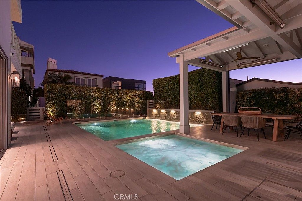 This luxurious outdoor living space features a modern swimming pool and integrated spa, illuminated by soft evening lighting. A covered patio area with a dining table provides a comfortable space for entertaining, while a tall, manicured hedge offers privacy. The scene is captured from a low-angle perspective, emphasizing the sleek wood-look decking and the tranquil, resort-like atmosphere.