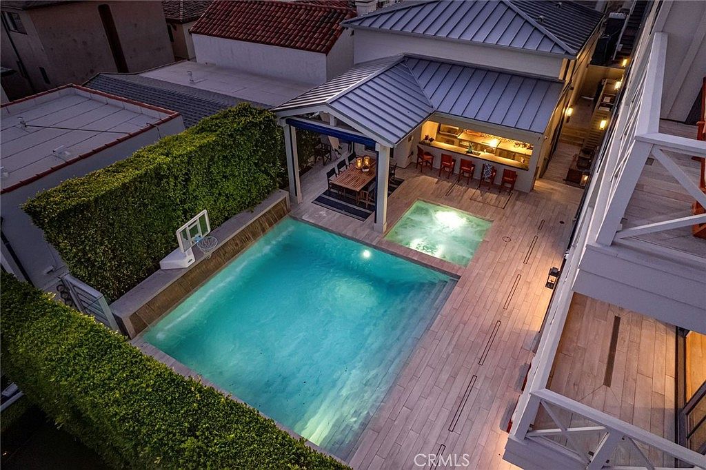 This high-angle aerial view captures a luxurious backyard oasis featuring a rectangular swimming pool and an adjacent square spa. A covered outdoor dining area with a wooden table and a built-in bar with stools provides a sophisticated space for entertaining. The area is enclosed by tall, manicured hedges and light-toned wood-look decking, creating a private and modern retreat.