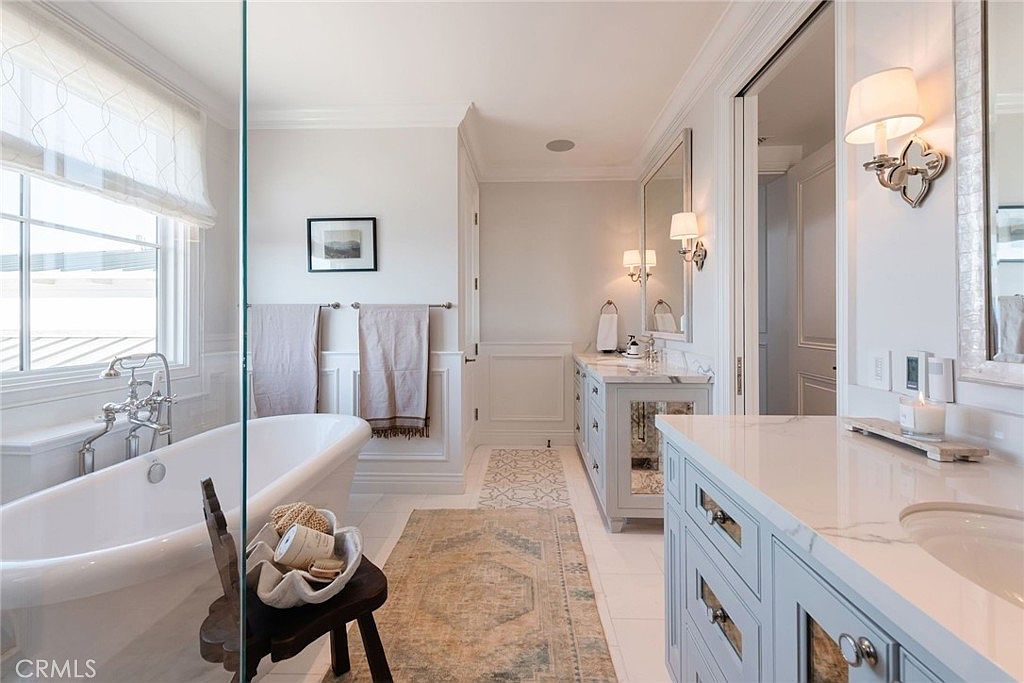 This elegant primary bathroom features a freestanding soaking tub positioned next to a large window with soft drapery. A double vanity with light-colored cabinetry and stone countertops sits opposite the tub, complemented by a patterned runner rug on the floor. The space is bright and airy, with a glass-enclosed shower area and sophisticated wall sconces providing a refined, spa-like atmosphere.