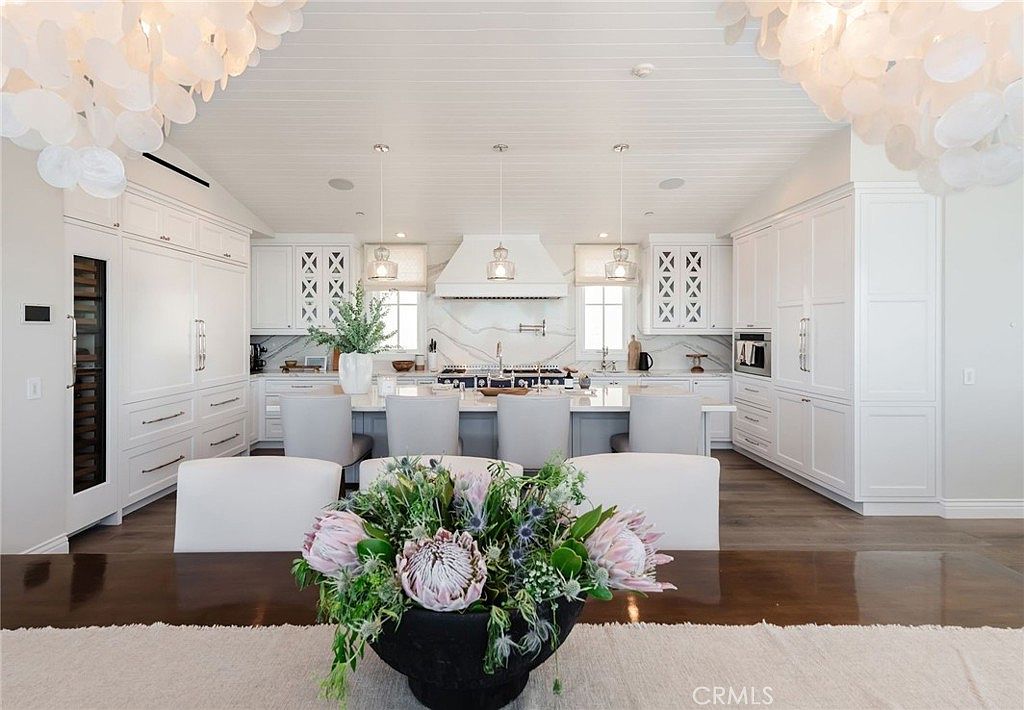 This bright and airy kitchen features a large central island with bar seating, white cabinetry, and a prominent stone backsplash behind the range. The space is illuminated by decorative pendant lights and large, ornate chandeliers, creating a sophisticated and open atmosphere. The perspective is captured from a dining area, looking directly into the kitchen, highlighting the clean lines and cohesive design.