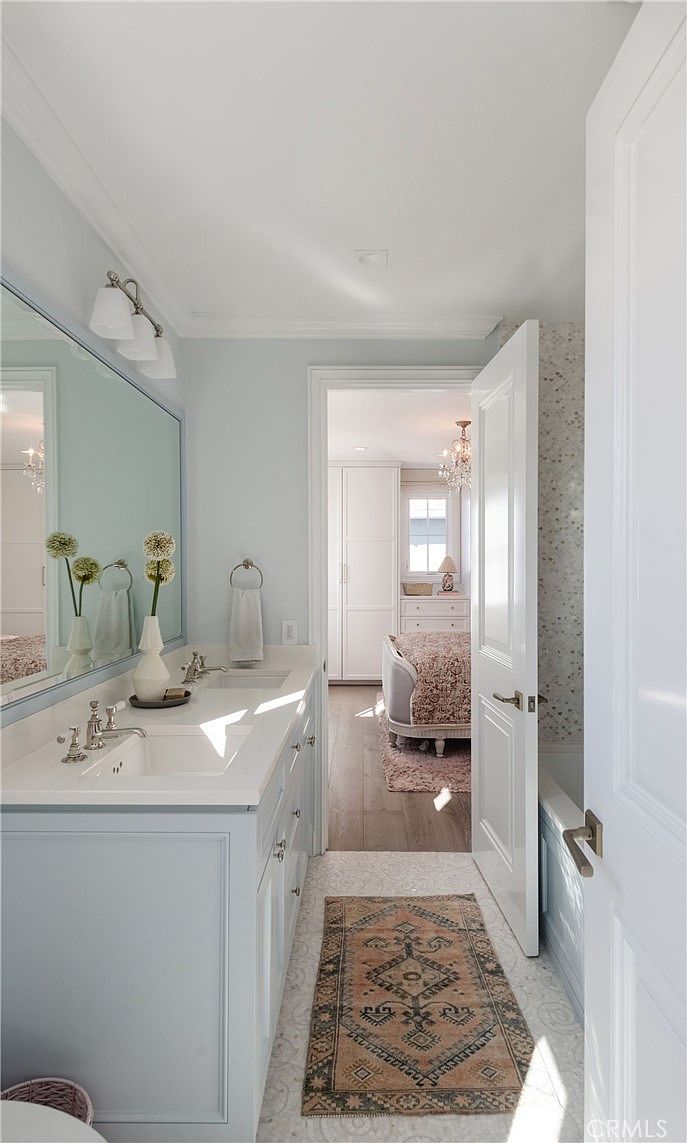This bright and airy primary bathroom features a double vanity with light blue cabinetry and a white countertop, complemented by a large mirror and elegant light fixtures. A patterned runner rug adds texture to the tiled floor, while an open door provides a direct view into an adjacent bedroom with a bed and dresser. The space is designed with a soft, cohesive color palette that creates a serene and inviting atmosphere.