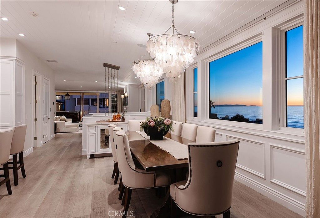 This elegant dining room features a long wooden table surrounded by upholstered chairs, positioned to take advantage of expansive ocean views through large windows. Two ornate, multi-tiered shell chandeliers hang above the table, adding a coastal-luxe aesthetic to the bright, white-walled space. The room flows seamlessly into an adjacent kitchen area, creating an open-concept layout that emphasizes natural light and a serene atmosphere.