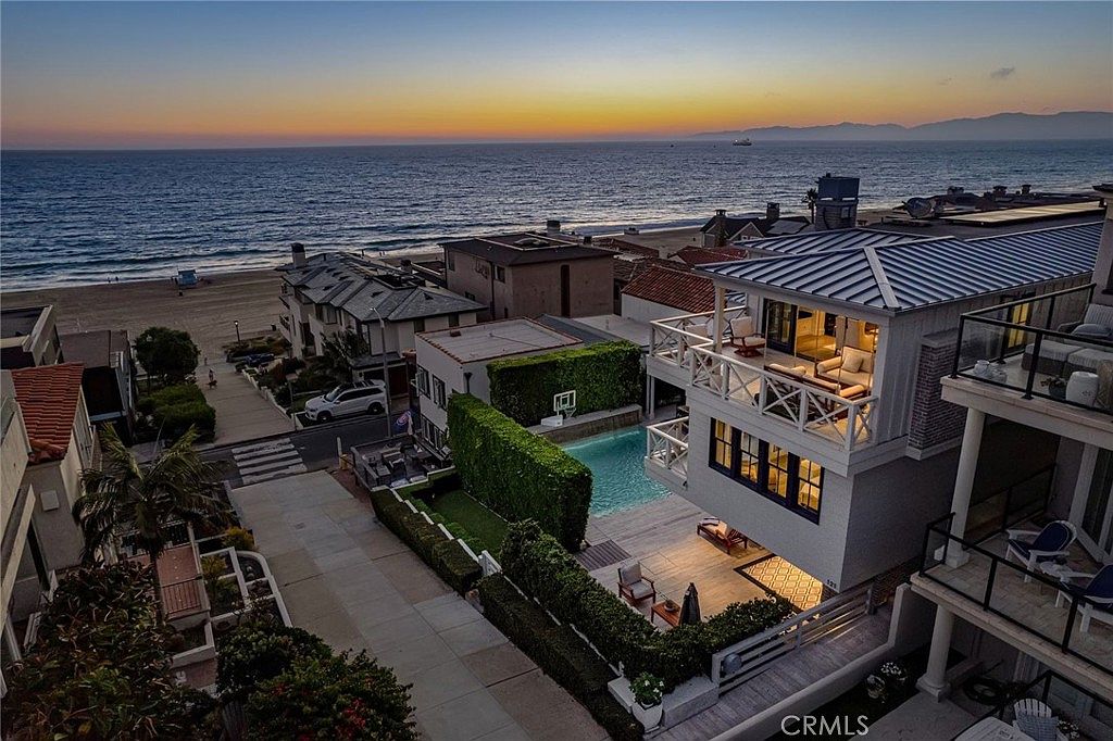 This high-angle aerial view captures a luxurious coastal property at sunset, showcasing its prime location steps from the beach. The home features a multi-level design with a private swimming pool, lush privacy hedges, and expansive balconies that overlook the ocean. The scene is bathed in the warm, golden light of dusk, highlighting the seamless transition between indoor living spaces and the outdoor environment.