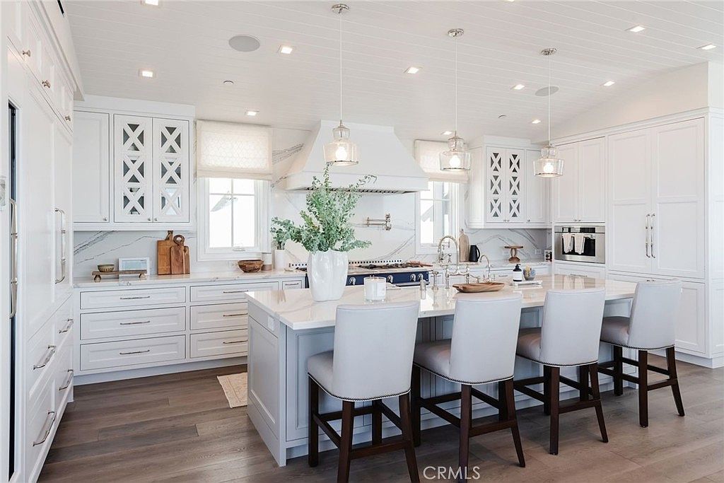 This bright and airy kitchen features a large central island with seating for four, topped with a light-toned stone surface. The space is defined by crisp white cabinetry, a custom range hood, and a marble-patterned backsplash, all illuminated by elegant pendant lighting and recessed ceiling lights. The perspective is a wide-angle shot from the kitchen island looking toward the range, showcasing a clean, sophisticated design with light wood flooring throughout.