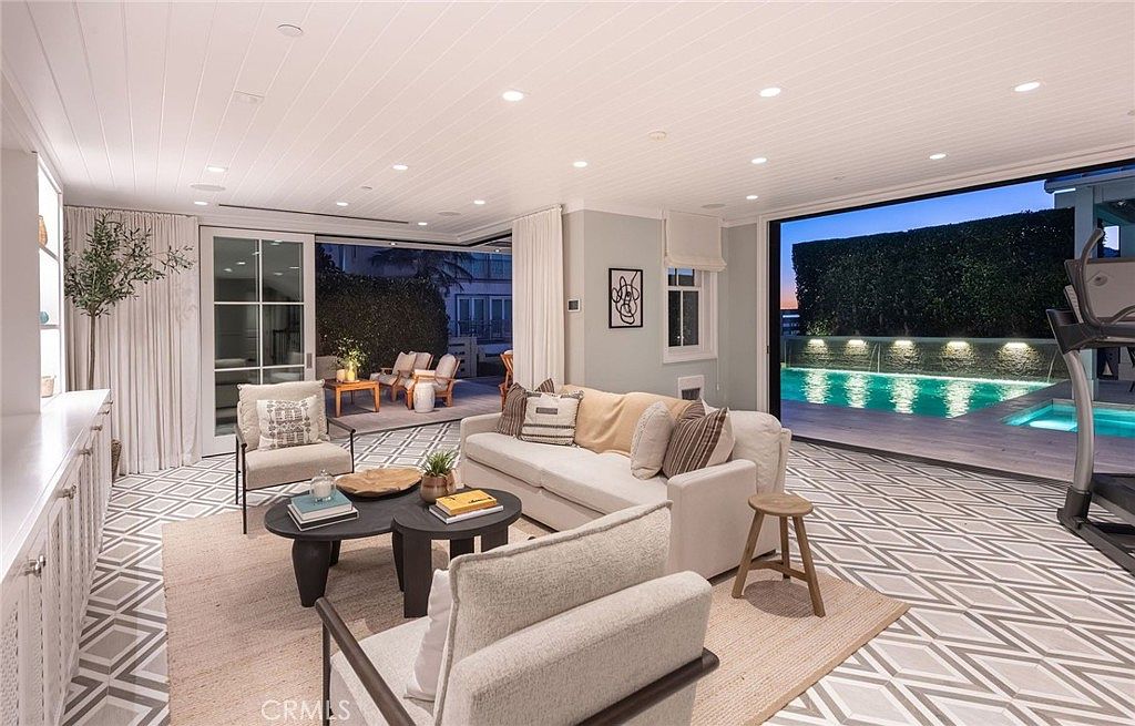 This bright and airy living room features an open-concept design that seamlessly transitions to an outdoor patio and pool area through large sliding glass doors. The space is furnished with a neutral-toned sofa, a pair of armchairs, and a tiered coffee table set, all arranged on a textured area rug. The room is characterized by its light-colored walls, white paneled ceiling, and a clean, modern aesthetic that emphasizes indoor-outdoor living.