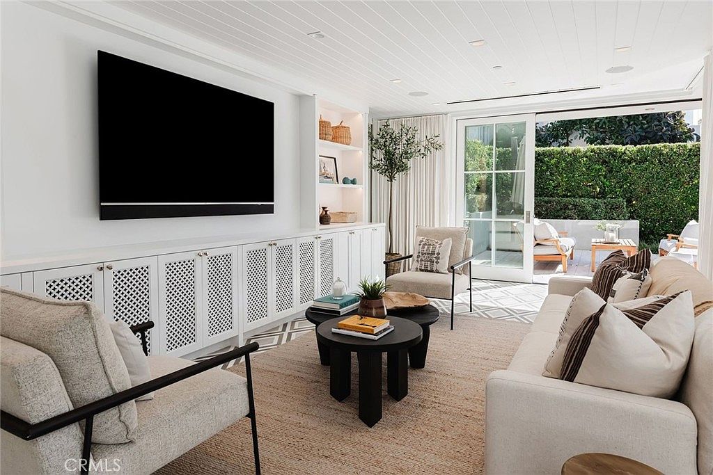 This bright and airy living room features a neutral color palette with textured furnishings, including a light-colored sofa and two accent chairs. A large television is mounted above a long, white media console with decorative cane-front cabinet doors, while a set of glass sliding doors opens to a private patio area. The space is anchored by a natural fiber rug and two round, dark-toned coffee tables, creating a sophisticated and inviting atmosphere.