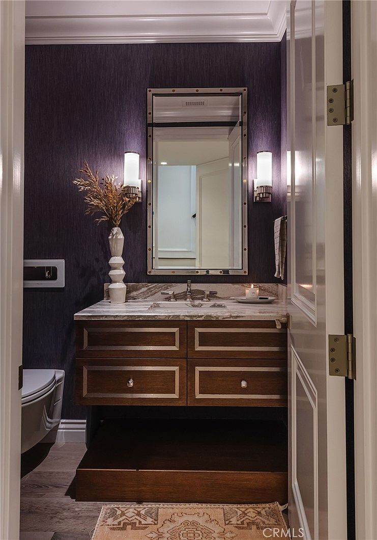 This elegant powder room features deep purple textured wallpaper that creates a sophisticated and moody atmosphere. The vanity is accented with metallic trim and topped with a stone counter, complemented by a rectangular mirror flanked by modern wall sconces. A small wooden step leads up to the vanity area, which is finished with a patterned rug on the floor.